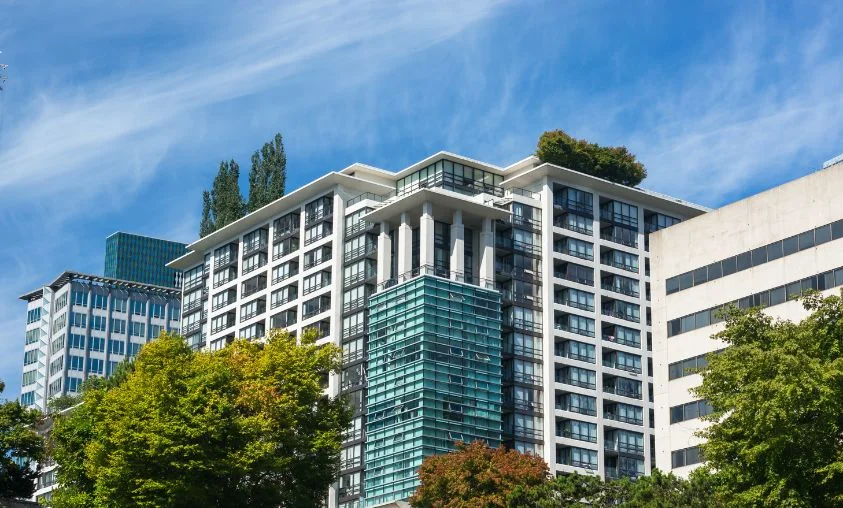 A modern high-rise building with glass and concrete architecture, featuring rooftop greenery and surrounded by trees under a clear blue sky.