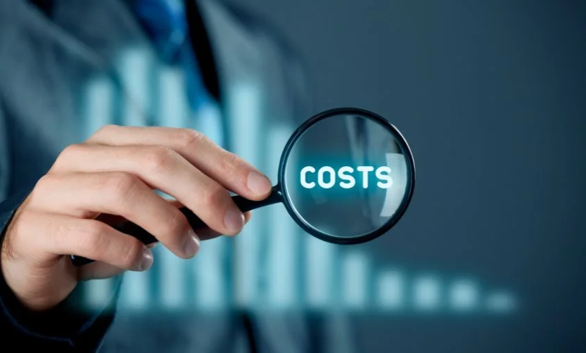 A person in business attire holding a magnifying glass over the word “COSTS,” with a blurred bar graph in the background, symbolizing financial analysis or cost management.